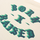 Close-up of a cream-colored t-shirt featuring a stylized graphic print of the words 'BORN TO BE' in a green and dark blue pixelated, zig-zag pattern.