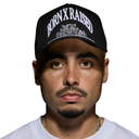 A man wearing a black baseball cap with white embroidered text reading 'BORN X RAISED' and a white graphic crest on the front, photographed against a plain white background.
