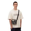 A model wearing a cream-colored t-shirt and dark jeans, carrying a small rectangular crossbody bag with a black strap and a green camouflage pattern on the front pocket, against a plain white background.