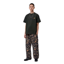 A male model stands against a plain white background wearing a dark olive green t-shirt with a small yellow and red chest graphic, paired with dark camouflage cargo pants and black shoes.