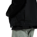 Close-up of a black jacket worn by a model, showing the sleeve, side pocket, and buttoned hem, paired with light gray pants against a white background.