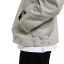 Close-up of a person wearing a light beige utility jacket over a white shirt and black pants, showing the sleeve, buttoned cuff, hem tab, and lower torso against a plain white background.