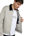 A man wearing a light beige zip-up jacket with a black collar and black interior lining, layered over a white graphic t-shirt, against a solid white background.