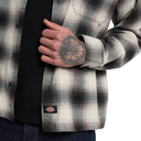 A close-up of a person wearing a Dickies plaid overshirt over a black t-shirt and blue jeans. The Dickies brand label is visible near the bottom left hem of the shirt.