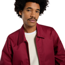 A man with dark curly hair and a mustache wearing a burgundy zip-up jacket over a cream ribbed sweater, photographed against a solid white background.