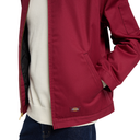 A close-up of a person wearing a burgundy zip-front jacket over a cream knit sweater and blue jeans. The jacket has a small multicolor Dickies logo patch near the lower hem and a black quilted lining visible on the left side.