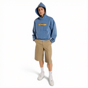 A model wearing a dusty blue pullover hoodie with a yellow and black 'Dixies' graphic on the chest, paired with tan shorts, white socks, and white sneakers, set against a plain white background.