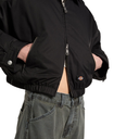 A close-up of a person wearing a black zip-front jacket with elastic cuffs and hem, paired with gray jeans, against a plain white background.