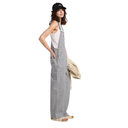 A woman modeling a pair of wide-leg gray striped corduroy bib overalls over a white tank top, holding a beige jacket, and wearing a black cap and beige sandals, set against a plain white background.