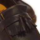 Close-up of a dark brown leather tassel loafer showing the pebbled texture, stitching, and double tassel detail against a white background.