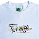 A close-up of a light blue t-shirt featuring a colorful graphic on the chest that reads 'Frog Skateboards' with cartoon frogs and a yellow star. The inside collar shows a green neck label with a green frog logo.