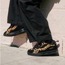 A person wearing black wide-leg pants and black low-top sneakers with a bold orange and black leopard print pattern, standing on concrete next to a light-colored brick wall.