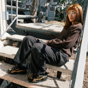 A person with strawberry-blonde hair and glasses sits on concrete outdoor stairs, wearing a brown knit sweater, dark cargo pants, and black sneakers with a yellow leopard print pattern.