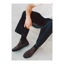 A person seated on a stool wearing dark brown suede Chelsea-style boots with black elastic side panels, black socks, and dark jeans on a light stone floor.