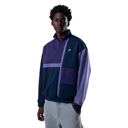 A man wearing a color-blocked quarter-zip sweatshirt with navy, purple, and lavender panels, paired with light gray jeans, against a plain white background.