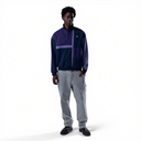 A model stands against a white studio background wearing a color-blocked quarter-zip pullover jacket in navy and purple, paired with light gray jeans and black shoes.