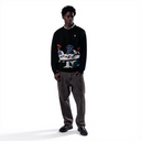 A man modeling a black crewneck sweater with a colorful graphic print on the front, paired with dark gray jeans and black shoes, against a solid white background.