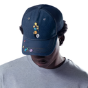 A dark navy blue trucker-style baseball cap with a cartoon dog graphic and colorful numbered pins on the brim, worn by a model in a gray t-shirt against a white background.