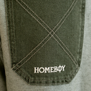 Close-up of an olive green denim pocket with diagonal stitching and a white embroidered 'HOMEBOY' logo on the front, set against a light gray fabric background.
