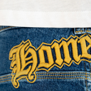 Close-up of blue denim jeans with a large yellow embroidered Gothic-style 'Home' patch on the back pocket, with a white shirt hem visible at the top.