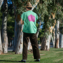 Person wearing a green t-shirt with a pink flower design and a straw hat, standing in a park.