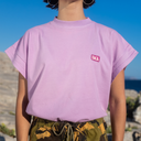 Person wearing a light purple t-shirt with a logo, standing outdoors against a blue sky.