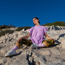 Person sitting on a rocky outcrop wearing a purple t-shirt and camouflage pants.