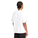A man with braided hair is shown from the back wearing an oversized white t-shirt and dark pants against a solid white background.