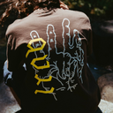 A person viewed from behind wearing a brown T-shirt with a large graphic print on the back. The design features a white hand outline and the word 'ROCK' in yellow gothic lettering.