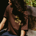 A person wearing a dark brown t-shirt with yellow text that reads 'here export your mix', photographed in an outdoor setting with warm sunlight and shadows.