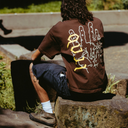 A person sitting outdoors on stone steps, viewed from behind, wearing a brown T-shirt with a large white and yellow graphic print on the back and dark shorts.