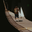 A person stands on a skateboard in a sunlit alley, viewed from behind, wearing a brown graphic T-shirt with a large yellow hand design and the word 'KOREA', dark shorts, white socks, and sneakers.