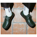 A person wearing dark green leather lace-up shoes with black soles, styled with white crew socks that read 'BEYOND FREEDOM' and black trousers, standing on terracotta tiled flooring.