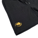 Close-up of a black garment, likely a hoodie or sweatshirt, showing a gold embroidered circular logo on the lower left leg. The fabric is textured, and a portion of a black drawstring is visible at the top against a plain white background.