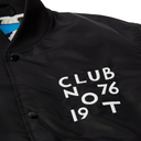 Close-up of a black jacket with white text reading 'CLUB 76 NOT 19 T' on the chest, plus black snap buttons and a ribbed collar.