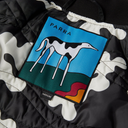 Close-up of a quilted jacket with a black and white camouflage pattern, featuring a colorful rectangular patch with a stylized white-and-black bird on a blue and green background and the word PARRA.