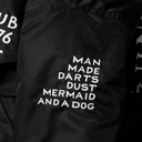Close-up of a black jacket or fabric panel with white printed text reading 'MAN MADE DARTS DUST MERMAID AND A DOG' and partial white lettering on the left and right edges.