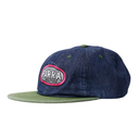 A dark blue denim snapback cap with an olive green flat brim and a circular embroidered patch on the front that reads 'PARRA SOLITUDE'.