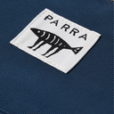 A close-up of a white rectangular Parra brand patch with a black fish skeleton logo and the word 'PARRA' stitched in black, set against a textured navy blue fabric background.