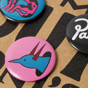 Three round pinback buttons displayed on a brown kraft paper background with black printed shapes. The main visible button is pink with a blue bird-like head illustration and red horns.