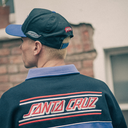 A person shown from behind wearing a Santa Cruz cap and a dark blue Santa Cruz sweatshirt with a light blue collar, set against an outdoor brick wall background.