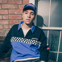 A man wearing a blue and black quarter-zip sweatshirt with a checkered stripe and 'DAVID CRUZ' text, paired with a matching blue cap, sitting in front of a brick wall and window.