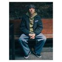 A person sitting on a wooden bench against a brick wall, wearing a black jacket over a green hoodie, blue jeans, and black sneakers. The jacket has a visible chest logo that appears to read Supreme.