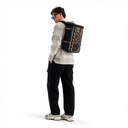 A model wearing a black and beige The North Face backpack, a beige and black plaid flannel shirt, black cargo pants, and sneakers, standing against a solid white background.