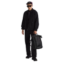 A model wearing a black jacket, black pants, black sunglasses, and a dark cap, holding a black backpack against a solid white background.