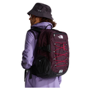 A person wearing a purple jacket and dark bucket hat shown from the back, carrying a large The North Face backpack in dark plum with bright pink laces on a white studio background.