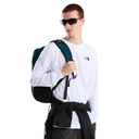 A man wearing black sunglasses, a white long-sleeve The North Face shirt, and a black jacket tied around his waist, carrying a dark green and black backpack against a solid white background.
