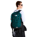 A person shown from the back wearing a teal and black The North Face backpack, a white long-sleeve shirt, and sunglasses against a plain white background.