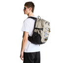 A man wearing a white t-shirt and black pants is shown from the back wearing a beige and black backpack with various outdoor gear attached to it, against a solid white background.