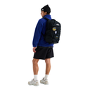 A man wearing a black beanie, blue fleece, black shorts, and white sneakers is shown from the back wearing a black The North Face backpack with multiple patches on a solid white background.